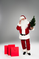 Santa claus talking on smartphone and holding small fir tree near shopping bags on grey background.