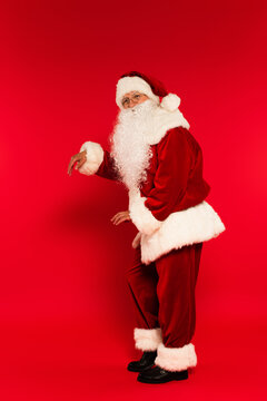Full length of bearded santa claus dancing and looking at camera on red background.