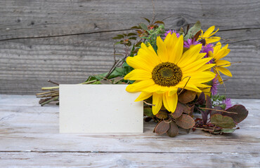 A bouquet of autumn flowers with a sunflower and a card with a copy space are on a wooden background.