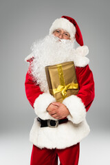 Santa claus in hat and eyeglasses holding present and looking at camera isolated on grey.