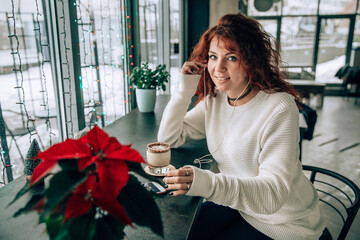 portrait of a young beautiful girl in a cafe with a cup of cocoa or coffee at Christmas time. Listen to music, enjoy and celebrate. Photo processing vintage and grain for mood and Christmas spirit