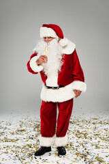 Santa claus in costume holding glass of champagne near confetti on grey background.
