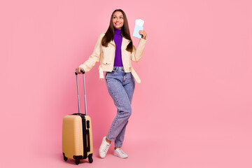 Full size photo of lovely millennial lady with valise hold tickets look promo wear jacket jeans sneakers isolated on pink color background