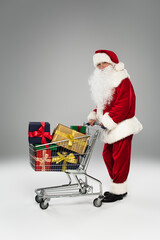 Full length of santa claus in costume and eyeglasses standing near shopping cart with gifts on grey background.