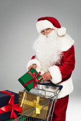 Santa claus in costume holding gift box near present in shopping cart isolated on grey.