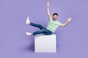 Full length photo of nice young guy sit white cube copyspace falling back wear trendy gray clothes isolated on violet color background