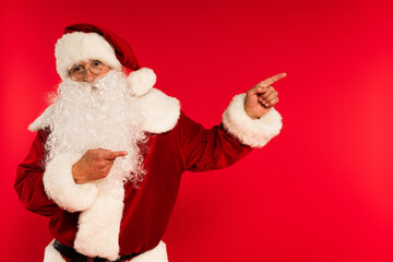 Santa claus in eyeglasses pointing with fingers and looking at camera isolated on red.