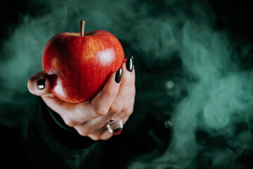 Woman as witch offers red apple as symbol of temptation, poison. Fairy tale, white snow wizard concept. Spooky halloween, cosplay. Smoke, haze background. - obrazy, fototapety, plakaty