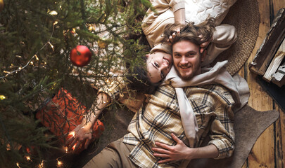 Hugging stylish love couple girl, guy lie on skin on floor in cozy wooden country house,chalet in winter forest.Celebrating new year eve.Gifts,family holiday.Romantic weekend.Decorated Christmas tree