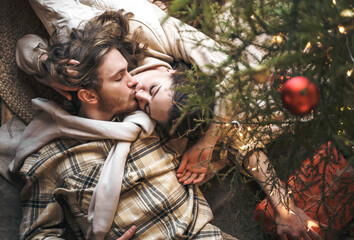 Kissing stylish love couple girl, guy lie on skin on floor in cozy wooden country house,chalet in winter forest.Celebrating new year eve.Gifts,family holiday.Romantic weekend.Decorated Christmas tree