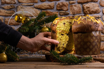 A hand taking a cut piece of Traditional Italian Christmas Cake Panettone with festive decoration on wooden rustic background. Artisan panettone is classic italian Christmas Food Edible gift
