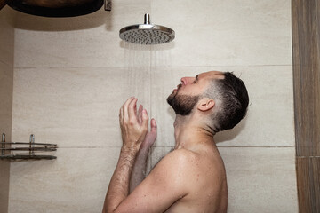 Young bearded man with tattoo taking shower in bathhouse (sauna) under water falling from rain shower head. Morning lifestyle guy showering. Concept body care hygiene. Copy text space