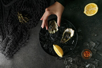 Woman hand hold a fresh opened oysters in a black plate with ice on black textured background.