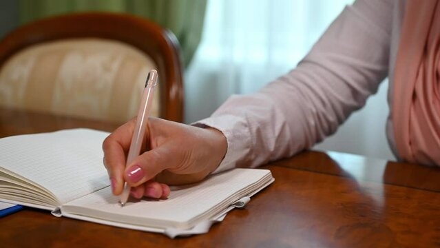 Close-up. Selective focus on the hand of woman in pink hijab, holding a pen and writing words in Arabic language, making notes on notepad while sitting at wooden table in a luxury interior