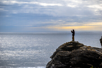 Hombre sobre una roca fotografiando el mar y el atardecer con un celular en día frío con pequeña luz naranja del sol al fondo detrás de la persona en pelluhue, chile