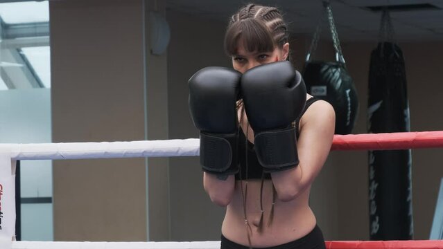 Female fighter with boxing gloves on her hands stands in a boxing ring leaning against the ropes.. Woman has an abrasion on her cheek.