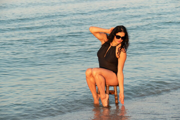 The girl is sitting on a stool in the sea. Brunette in a black swimsuit on the beach.