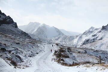 Snow-covered highland gorge with rocky peaks, a glacier and a road, two people are walking along the road, morning, the sun is shining, the sky is in the clouds