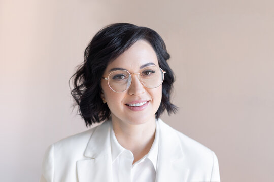 Portrait Of A Smiling Middle-aged Brunette In A White Jacket And Trendy Round Glasses. Business Woman On A Plain Background Of Cocoa Color.