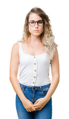 Beautiful young blonde woman wearing glasses over isolated background with serious expression on...