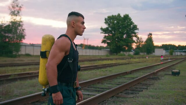 Portrait of a handsome muscular man in a black tank top with a dragger on his back who stands on the railway tracks at sunset