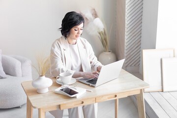 Smiling brunette woman in a white jacket and trousers. Works on a laptop while sitting at a wooden table in a minimalist interior by the window. typing text in online chat.