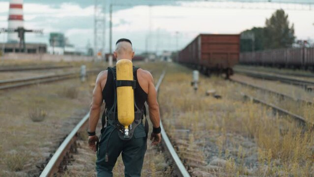 Young muscular man with balloon dragger on the back goes along the rails of marshalling yard near an industrial area and looks back