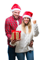 Young couple in love wearing christmas hat and holding present over isolated background annoyed and frustrated shouting with anger, crazy and yelling with raised hand, anger concept