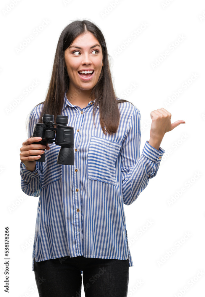 Sticker Young hispanic woman holding binoculars pointing and showing with thumb up to the side with happy face smiling