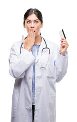 Young hispanic doctor woman holding credit card cover mouth with hand shocked with shame for mistake, expression of fear, scared in silence, secret concept