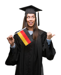 Young hispanic woman wearing graduated uniform holding flag of germany pointing and showing with thumb up to the side with happy face smiling