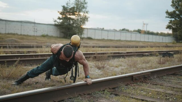 Young muscular man with balloon drager on his back is training at railway yard and doing push-ups from the rails on one arm