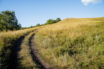 Dirt road steeply rises up the field, mountainous terrain, evening landscape, smooth turn of the road, horizon line, at the top of the mountain, Russian field