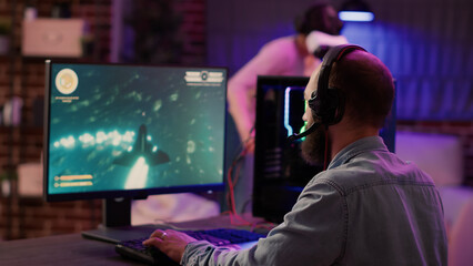 Over shoulder view of gamer playing multiplayer space shooter while gaming girl is fighting in virtual reality in home living room. Man streaming action game on pc while gaming girl uses vr goggles. © DC Studio