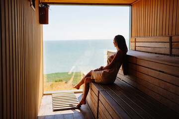 woman in a towel sits on a bench in the sauna with the panoramic window. 