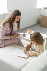 Cheerful female psychologist holds a meeting with a little boy through games and drawing. Makes notes. Light office.
