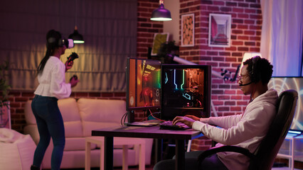 Gamer playing multiplayer first person shooter while gaming girl is fighting in virtual reality boxing game. Man streaming online action game on pc while girlfriend uses vr goggles for simulation.