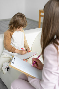 Little boy psychologist's appointment. He draws  picture of his family request of  psychologist. An unrecognizable psychologist analyzes drawing and makes notes for himself. Blurred foreground.