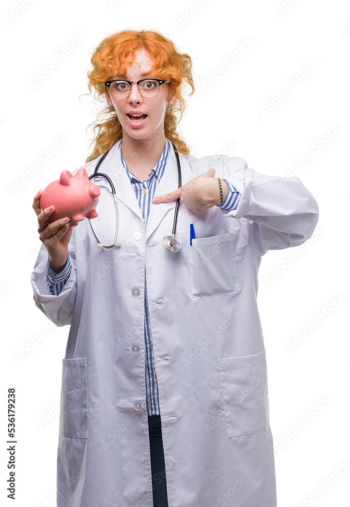 Wall mural young redhead doctor woman holding piggy bank with surprise face pointing finger to himself