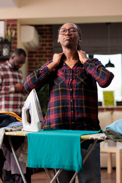 Stressed African American Woman Does Domestic Chores While Distracted Male Roommate Uses Cellphone. Exhausted Frustrated Modern Woman Finishes Ironing Clean Clothes In Urban Apartment