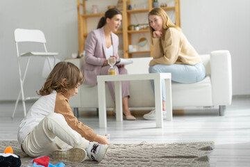 Mother with a minor son at a reception with a child psychodog. The son is sitting on a blanket on the floor playing with toys. Spacious office.