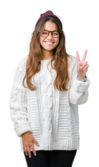 Young beautiful brunette hipster woman wearing glasses and winter hat over isolated background showing and pointing up with fingers number two while smiling confident and happy.