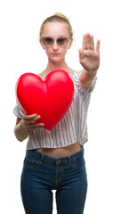 Blonde teenager woman holding red heart with open hand doing stop sign with serious and confident expression, defense gesture