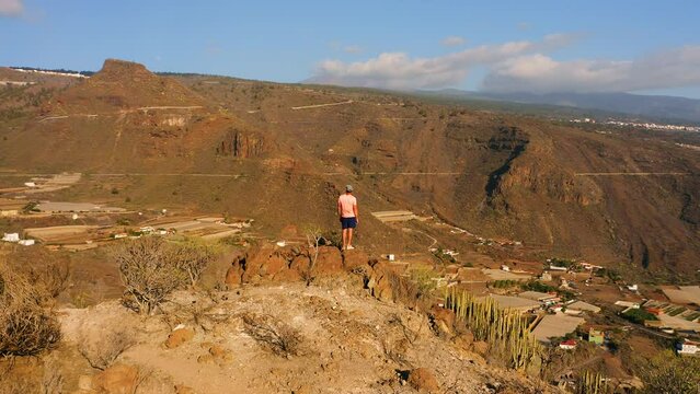 Tenerife Canary Island, mountain landscape. Tourist man at the edge of the sheer cliffs. Hiking area. Amazing nature, scenery. Village Tamaimo in a gorge. Drone circular flight around a person.