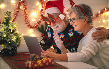 Happy senior couple wearing Santa hat at Christmas time in video call by laptop from home getting in touch with family or friends