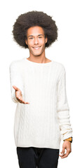 Young african american man with afro hair wearing winter sweater smiling friendly offering handshake as greeting and welcoming. Successful business.