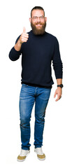 Young blond man wearing glasses and turtleneck sweater doing happy thumbs up gesture with hand. Approving expression looking at the camera showing success.