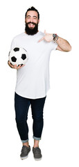 Young man with long hair and bear holding soccer football ball with surprise face pointing finger to himself