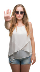 Young blonde woman wearing sunglasses doing stop sing with palm of the hand. Warning expression with negative and serious gesture on the face.