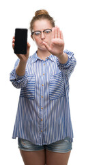 Young blonde woman using smartphone with open hand doing stop sign with serious and confident expression, defense gesture